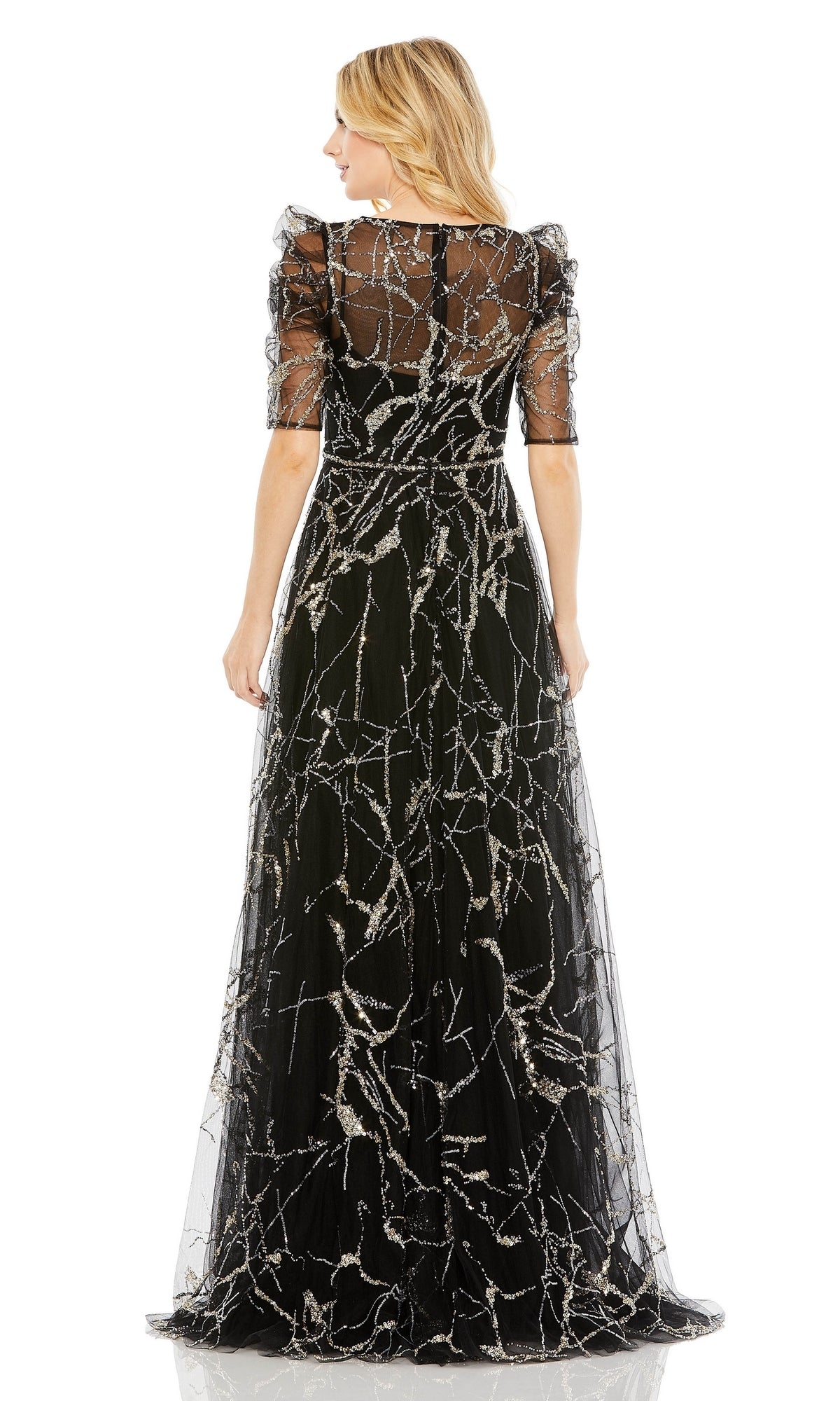 A woman stands facing away, wearing the Long Formal Dress 20428 by Mac Duggal—a black A-line gown with sheer sleeves, a beaded belt, and intricate silver branch-like patterns.